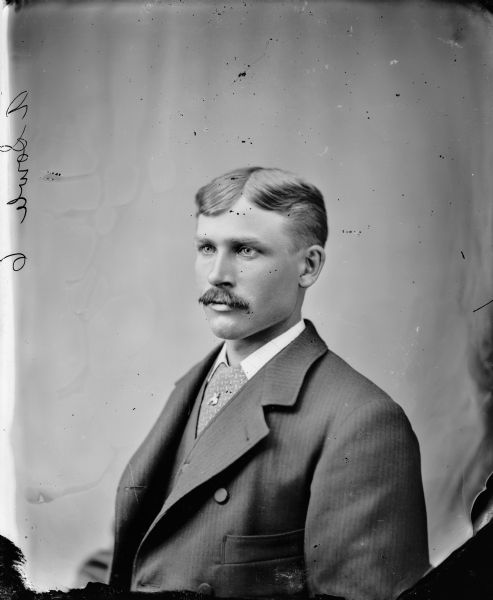 Waist-up studio portrait of a European American man, A. Sowle, with a moustache. He is posed sitting wearing a dark-colored suit coat, wide necktie, and tie pin. Etched on the glass plate negative is, "A. Sowle 6."