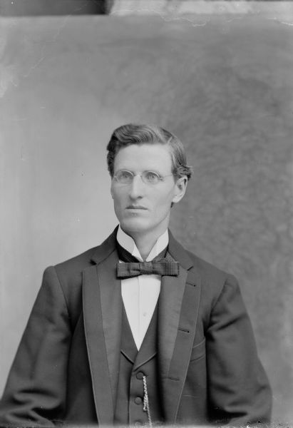 Waist-up studio portrait of a European American man wearing eyeglasses posed sitting and wearing a dark-colored suit coat, bow tie, and chain watch fob. Man identified as Hans Johnson.