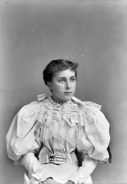 Studio portrait of a young European American woman posed sitting and wearing a light-colored dress with puffed sleeves, ribbons, belt, and a ruffled collar and bodice trimmed with lace. Woman identified as Grace Ogden.
