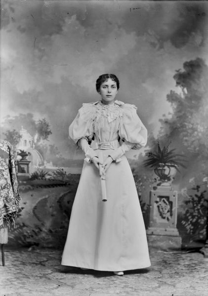 Studio full-length portrait of a European American woman posed standing and holding a diploma in front of her with both hands. She wears gloves and a light-colored dress with puffed sleeves, ribbons, belt, and a ruffled collar and bodice trimmed with lace. Woman identified as probably Grace Ogden.