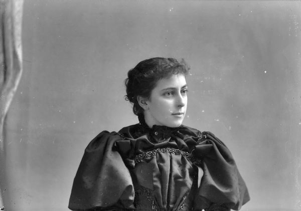 Waist-up studio portrait of a young European American woman posed sitting and wearing a dark-colored dress with puffed sleeves and beaded trim on the bodice. Woman identified as Grace Ogden.