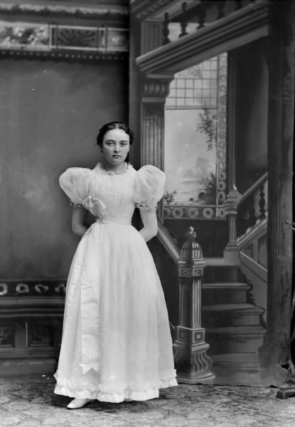 Studio full-length portrait in front of a painted backdrop of a European American woman posed standing with her arms folded behind her. She is wearing a light-colored dress with short puffy sleeves. There is a large bow at her waist which reaches to the hem of the dress. There are small flower decorations around the neck collar and bottom of the sleeves. She appears to be wearing long gloves. Woman identified as Ella Stiehl.