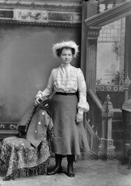 Studio full-length portrait in front of a painted backdrop of European American woman posed standing with her right arm resting on the back of a chair draped with a dark-colored coat over a tasseled cloth. She wears a dark-colored skirt, light-colored ruffled blouse, and a feathered hat. Woman identified as probably Frieda Schoesser.