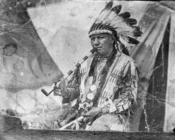 Copy photograph of a Ho-Chunk man posing sitting outside a tent, smoking a calumet pipe, holding a metal tomahawk, and wearing a Sioux headdress, several necklaces, and gorgets. He is identified as John Buffalo Head.