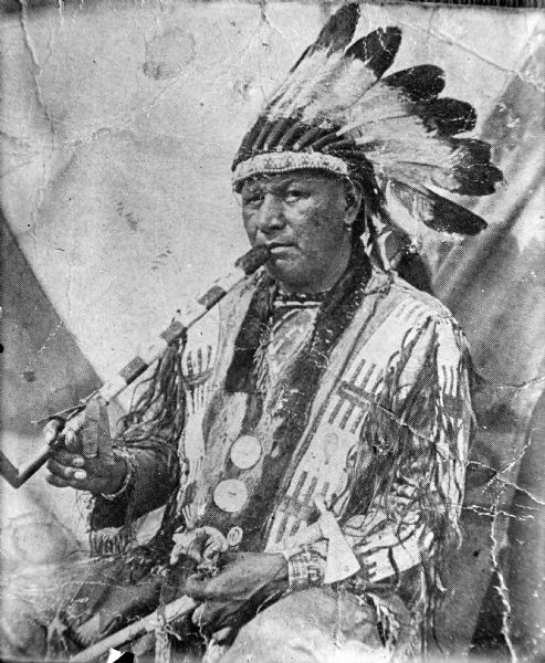 Copy photograph of a Ho-Chunk man posing sitting outside a tent smoking a calumet pipe, holding a metal tomahawk, and wearing a Sioux headdress, several necklaces, and gorgets. He is identified as John Buffalo Head.