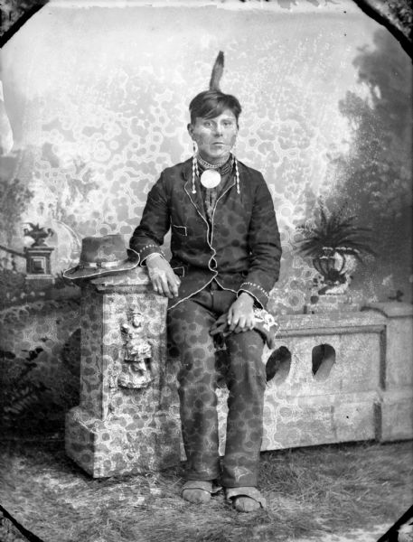 Full-length studio portrait in front of a painted backdrop of a Ho-Chunk man posing sitting on a prop stone wall. His hat, which has a German silver hat band, is sitting on the stone column, and he is wearing a beaded coat, moccasins, gorget, Canadian 5 cent coin chain earrings, and an erect eagle feather (symbolizing war honor). He is identified as Jasper Blowsnake.