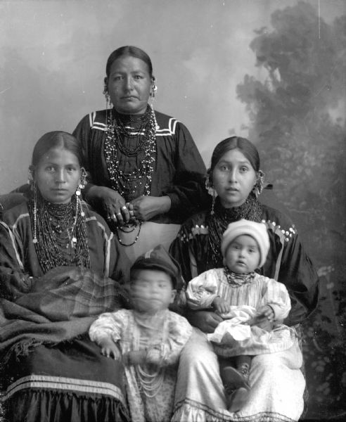 Studio portrait of Ho-Chunk women and children. All the women are wearing several necklaces and earrings, and the child in the center front has moved and his face is blurred. In the background is a painted backdrop. Names (standing) Ruby Lowcloud, Otter. (Sitting, l to r) Emma Thunder, Littlesoldier (We Pa Ma Ka Ra Win Kah) (Daughter of [Wa Con Cha Kah] John Thunder aka Dr. Thunder and [We Hon Pe Kaw] Lucy Bear, Thunder), (standing, center, blurred) Emma's son, Simon Robert Littlesoldier (Wo No Ka Re Hunk Kah) (Son of David Bow Littlesoldier and Emma Thunder, Littlesoldier), Lena Ora Longthunder, Stacy and her baby, Alice Hilda Stacy, Funmaker.
