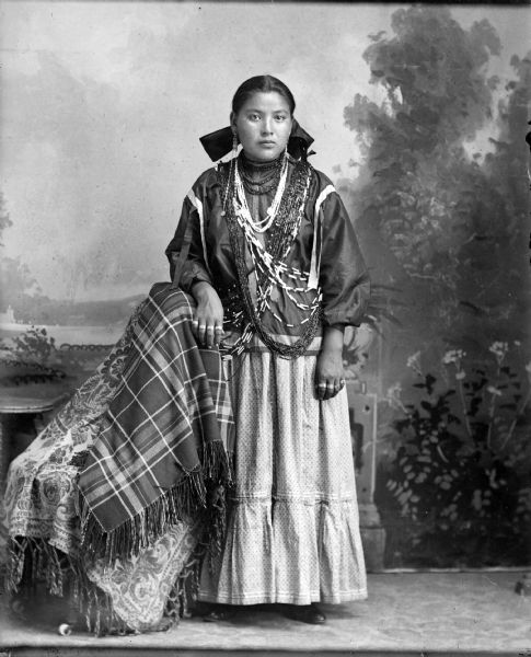 Studio full-length portrait in front of a painted backdrop of a young Ho-Chunk woman posing standing. She has her right arm resting on a cloth covered chair. She is wearing several bead bandoleers, necklaces, earrings, and rings.