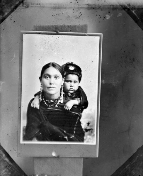 Copy photograph of a waist-up studio portrait of a woman in Ho-Chunk regalia posing sitting, wrapped in a blanket, with a child on her back. She is wearing long earrings and a necklace, and the child is wearing a fur hat. They are identified as Clara Kingsley Blackhawk and her son, Andrew who was a World War I veteran.