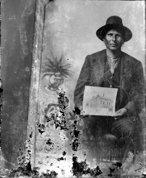 Copy photograph of a studio portrait of a Ho-Chunk man posing sitting in front of a painted backdrop, holding a photograph of a house, and wearing a suit and hat. Identified as Jim Green Grass, or Old Greengrass, who was Eli Youngthunder's grandfather.