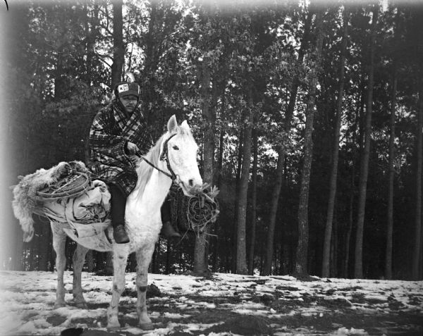 A Ho-Chunk man, identified as David Goodvillage, is wrapped in a plaid blanket and wearing a hat. He is posing mounted on a white horse loaded with hunting and trapping gear, including snowshoes. He is on a hill in front of trees, and snow is on the ground.