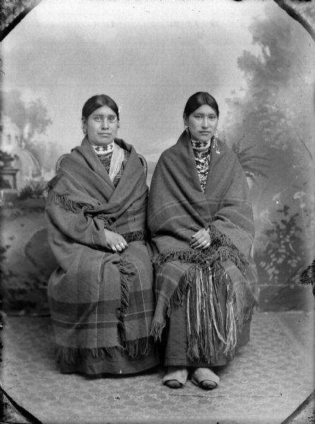 Studio portrait of two Ho-Chunk women posing sitting in front of a painted backdrop. They are both wearing several necklaces, earrings, beaded collars, and are wrapped in plaid fringed shawls. The woman on the left is identified as (?) Johnson.