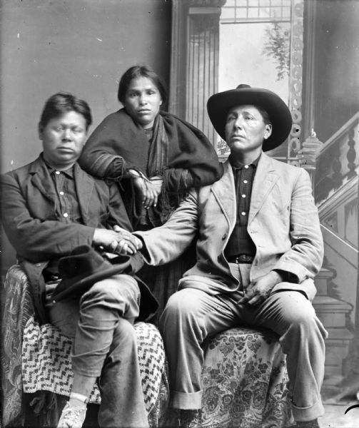 Studio portrait of two Ho-Chunk men posing sitting in front of a painted backdrop with their right hands clasped. Both men are wearing suit coats, and the man on the right is wearing a hat. The face of the man on the left is blurred by movement, and he has a hat on his lap. A Ho-Chunk woman is posing standing between the men behind their chairs. She is wrapped in a fringed shawl. The woman is identified as the wife of Will Decorah.