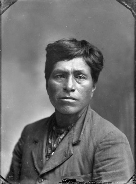 Quarter-length studio portrait of a Ho-Chunk man with short hair posing sitting. He is wearing a suit coat and plaid shirt. Identified as the father of Ben Decorah.