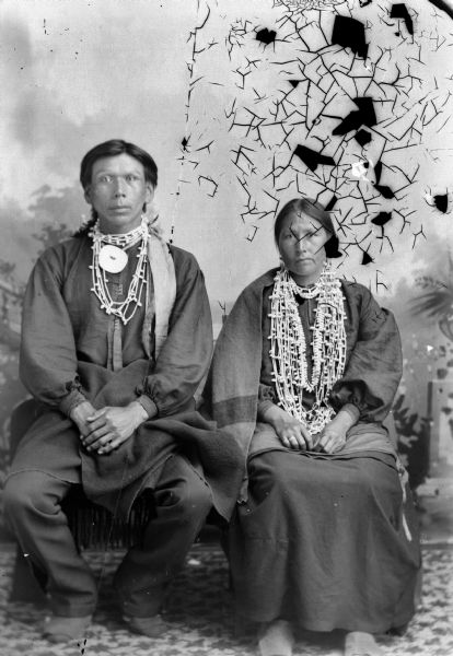 Full-length studio portrait of a Ho-Chunk man and woman, Mr. and Mrs. Luke Eagle, posinh sitting in front of a painted backdrop. The man on the left is wearing a few necklaces, a gorget, and a blanket around his waist, and the woman on the right is wearing several necklaces, earrings, a ring, and a shawl/blanket over her shoulders. Mrs. Luke Eagle is the sister of John Hazen Hill.