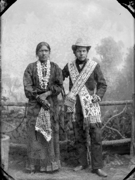 Full-length studio portrait of a Ho-Chunk woman and man posing standing in front of a prop wooden fence and painted backdrop. The woman on the left has her left hand holding her right forearm, and she is holding a handkerchief. She is wearing several necklaces and earrings. The man on the right is wearing Ho-Chunk bandoleers, earrings, a suit, and a hat. They are identified as Henry Rice Hill and his wife.