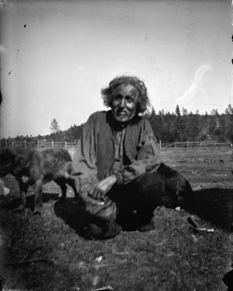 An elderly Ho-Chunk man with a beard is posing sitting in a field with two dogs. He is identified as Nehamakeewakh (Slits His Own Throat). A fence and trees are in the background. There is a hammer on the ground next to him.