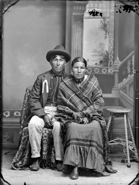 Full-length studio portrait of a Ho-Chunk man and woman posing sitting in front of a painted backdrop. The man on the left is wearing a suit coat, beaded shirt, pin with ribbons, and hat. The woman on the right is wearing several necklaces, and earrings, and is wrapped in a plaid fringed shawl. Identified as Charles Smith and his wife.