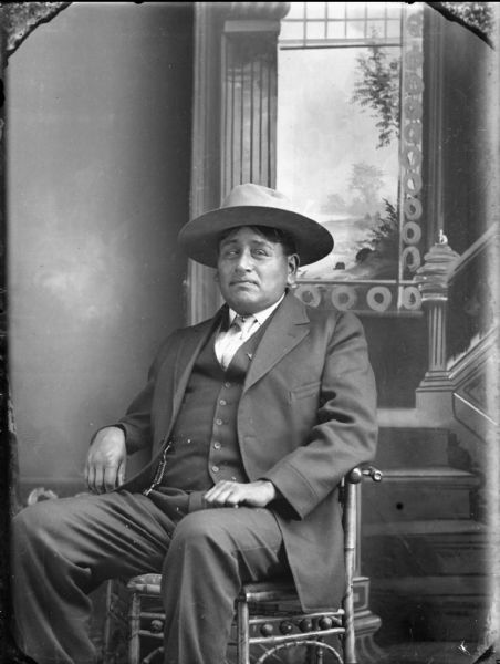 Studio portrait of a Ho-Chunk man posing sitting in front of a painted backdrop, wearing a suit and hat. He is identified as Schawinka Snyder Bear of Thurston, Nebraska.