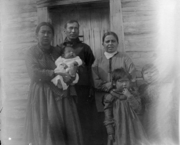 A Ho-Chunk man, two women, two children, and an infant are posing standing in front of a wooden building near the door. They are identified as probably John Stacy and his family.