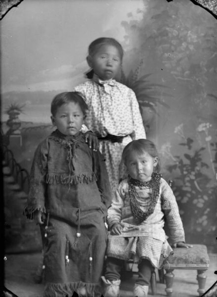 Studio portrait of a Ho-Chunk boy and girl posing sitting in front of a Ho-Chunk girl posing standing, all in front of a painted backdrop. The boy sitting on the left is wearing a beaded buckskin blouse and pants, the girl sitting on the right is wearing a light-colored dress, moccasins, and several necklaces, and the girl posing standing is wearing a light-colored print dress. They are identified as the Lewis children, George, Emma, and Mary, although not identified by position.