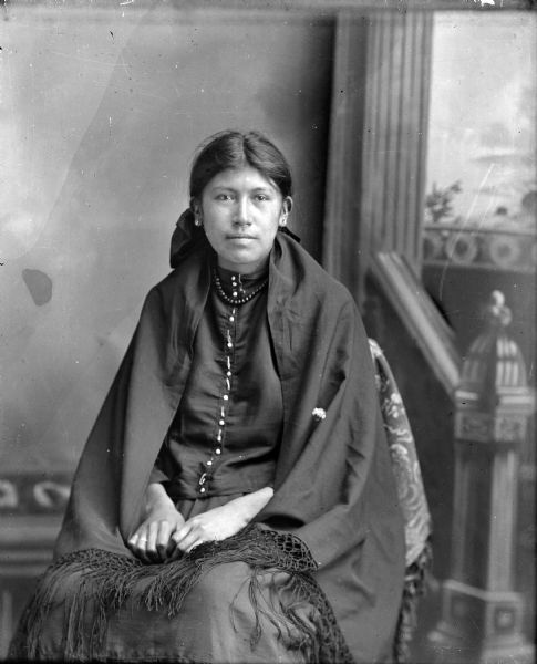 Studio portrait of a Ho-Chunk woman posing sitting in front of a painted backdrop. She is wearing a dark-colored beaded shirt, necklace, stud earrings, and a fringed shawl, and is identified as Annie Lincoln.