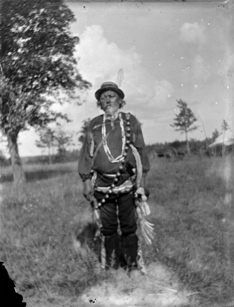 Outdoor portrait of an elderly Ho-Chunk man with a beard posing standing in a field wearing several necklaces, long bandoleer, and hat. He is identified as Old Little Soldier (Nojinka, Strikes Tree) of the Thunder clan, the father of Sam Little Soldier, and reportedly a Civil War scout.
