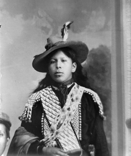Copy photograph of a waist-up studio portrait of a young Ho-Chunk man with long hair posing standing in front of a painted backdrop. He is holding a shawl over his right forearm, and wearing palm and woodland bandoleers, a beaded shirt, and a hat with an eagle feather. He is identified as David Little Soldier. There is a portion of a young man's face in the bottom left corner of the image that is cut off.