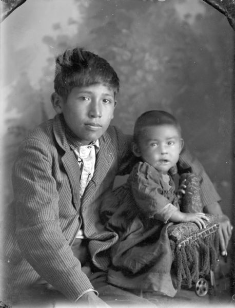Studio portrait of a Ho-Chunk boy posing sitting in front of a painted backdrop wearing a striped suit. He has his left arm around a Ho-Chunk infant wearing a long dress and perched on the arm of a chair. The boy is identified as Sam Lowe.