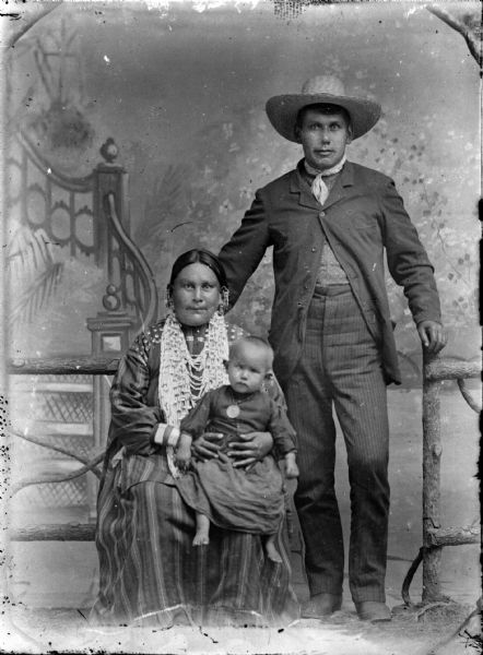 Studio portrait of a Ho-Chunk woman wearing several necklaces and earrings, sitting on the left. She is holding a Ho-Chunk child wearing a gorget. A Ho-Chunk man is standing on the right wearing a suit, bandana, and hat. They are posing near a prop wooden fence in front of a painted backdrop. Identified as Melvin Miner and his wife, Kate Thunder Miner.