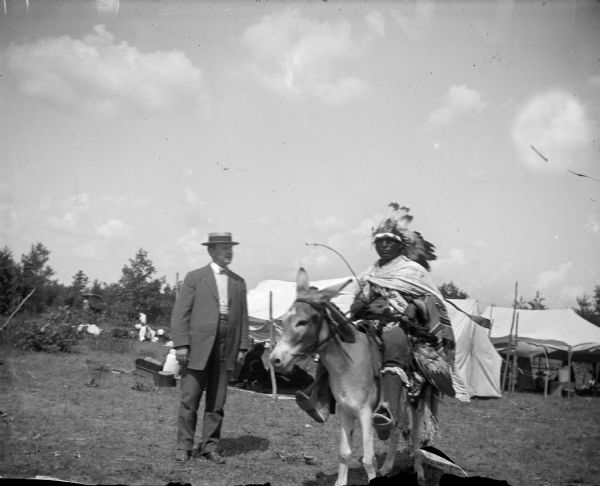 A Ho-Chunk man on the right is sitting on a mule wearing a Sioux headdress along with other regalia, in a field in front of tents and trees. A European American man is posing standing on the left nearby, and who is wearing a suit and straw hat. There are also people in the background in the tents. The Ho-Chunk man is identified as George Monegar.