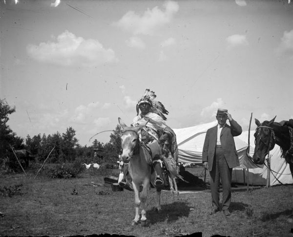 A Ho-Chunk man on the left is posing mounted on a mule, and is wearing a Sioux headdress among other regalia. A European American man is posing standing on the right wearing a suit and straw hat. Both are in a field in front of tents and trees. There is a horse on the far right, and a carriage in the far background on the left. The Ho-Chunk man is identified as George Monegar.