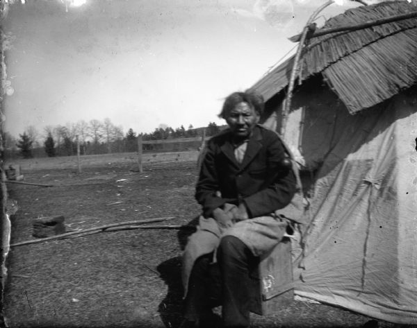 Elderly Ho-Chunk man, identified as Frank Mike, posing sitting outside a lodge wearing a suit coat and a shawl wrapped around his waist. In the background is a wooden fence and trees.