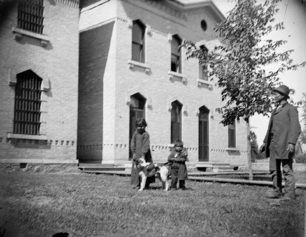 A Ho-Chunk man, two children, and a dog are standing outside a large brick building with bars over the windows. Identified as probably John Mike and the children of Jim Swallow at the County Jail. Jim Swallow was in jail for killing George Black Hawk in 1895.