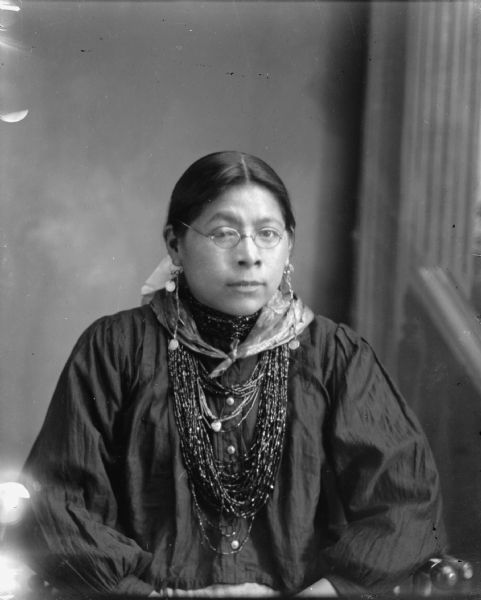 Waist-up studio portrait in front of a painted backdrop of a Ho-Chunk woman posing sitting and wearing several necklaces, a bandana, earrings, and eyeglasses. She is identified as Kate Massey(Massie), the daughter of William Massey and Jane Winneshiek (the oldest sister of Nellie Red Cloud).