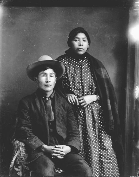 Studio portrait of a Nebraskan Winnebago man sitting on the left and wearing a suit, short tie, and hat, and a Ho-Chunk woman standing on the right wearing a polka dot dark-colored dress, a ring, and a fringed shawl. They are identified as James Priest and Kate Massey. They are Annie Massey's parents. In the background is a painted backdrop.