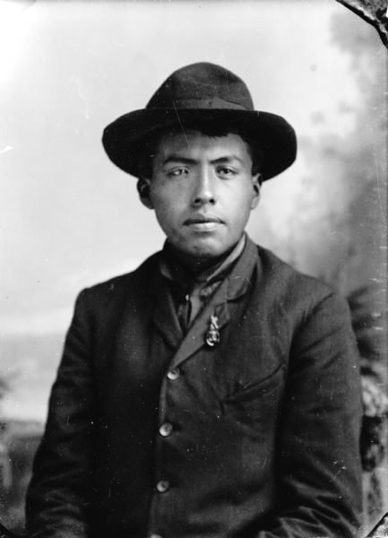 Waist-up studio portrait in front of a painted backdrop of a young Ho-Chunk man posing sitting and wearing a dark-colored suit, photograph button, bandana, and hat. He is identified as the son of Nose of Bear Wiggles (Pojorosopka) of the Bear Clan.