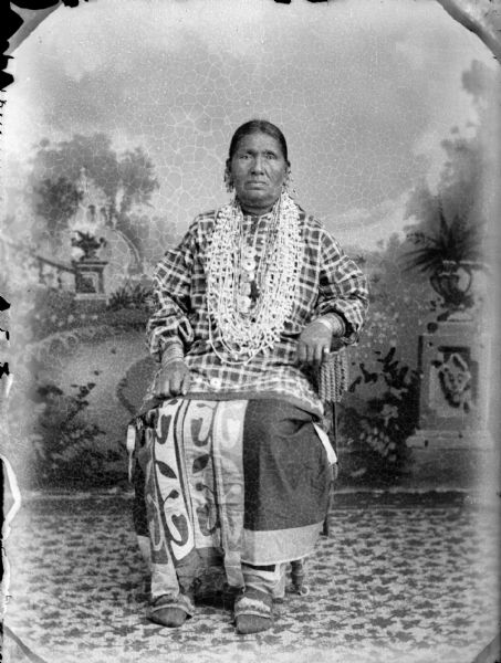 Studio portrait of a Ho-Chunk woman posing sitting in front of a painted backdrop wearing several necklaces, earrings, file bracelets, and a ribbon work skirt. She is identified as Betsy Thunder.
