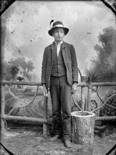 Full-length studio portrait of a young Ho-Chunk man posing standing in front of a prop wooden fence and a stump in front of a painted backdrop. He is wearing a suit, bandana, earrings, and a hat with an eagle feather. Identified as John Thunder.