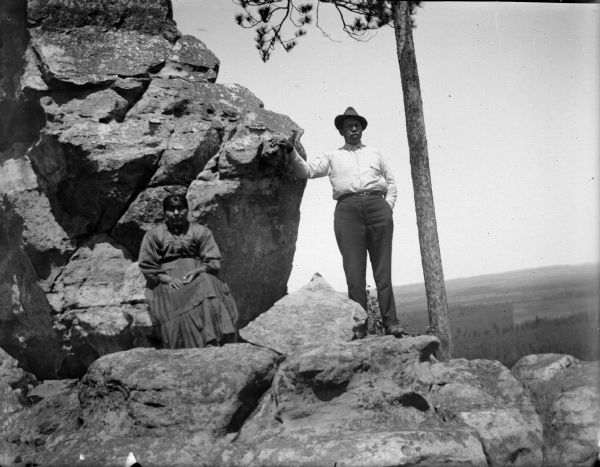 View of a Ho-Chunk man posing standing, and a Ho-Chunk woman posing sitting on a rocky outcrop near a tree. Far below in the background are fields, hills, and trees. Identified as probably Adam Thunder Cloud and his wife at or upon Old Stony (Xetenidi), near Shamrock.