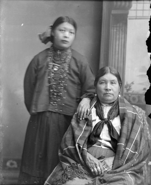 Studio portrait in front of a painted backdrop of a Ho-Chunk woman, who is wearing several necklaces and a hair ribbon, posing standing on the left with her right hand behind her back and her left hand on the right shoulder of another Ho-Chunk woman who is sitting. The woman on the right is wearing several rings, a bandana, earrings, and a shawl over her shoulders. The woman on the left is identified as Mary White Swan whose husband was Dr. White. The woman on the right is possibly a relative visiting from Nebraska.