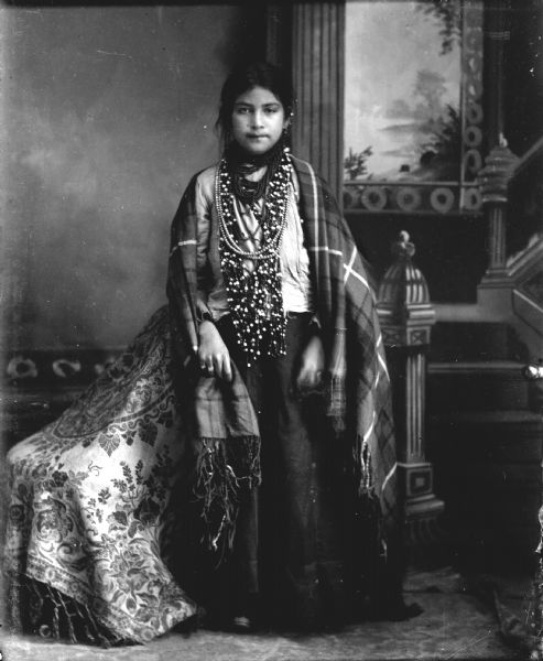 Studio full-length portrait of a Ho-Chunk girl posing standing near a chair, in front of a painted backdrop. She is wearing several necklaces, as well as earrings, rings, and a shawl draped over her shoulders. She is identified as Fanny Winneshiek, who never married because she died young. She was the sister of Ellen Winneshiek Garvins, the wife of George Garvins.