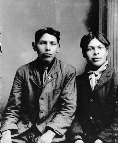 Waist-up studio portrait of two young Ho-Chunk men posing sitting in front of a painted backdrop. The men are both wearing suits and bandanas. The young man on the right is identified as Charles Young Thunder.