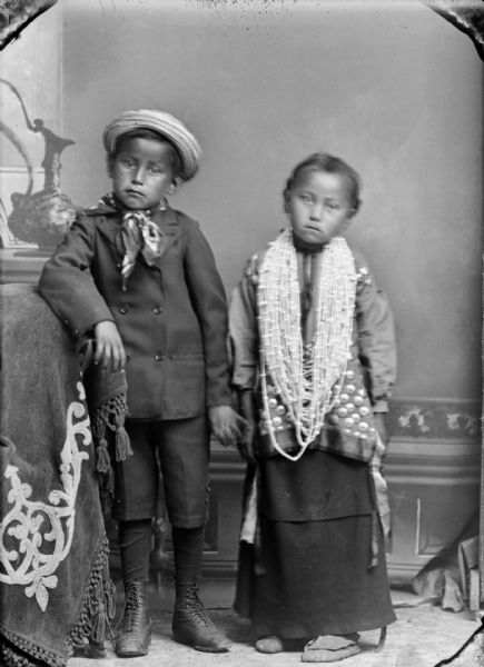 Studio full-length portrait in front of a painted backdrop of a young Ho-Chunk boy posing standing on the left, wearing a winter coat, knickers, bandana, boots, and a hat. On the right is a Ho-Chunk girl posing standing who is wearing a traditional blouse, moccasins, earrings, and several necklaces. They are identified as Stephen and Gladys Waukon. The boy's right arm is resting on a piece of furniture covered with cloth, and his left hand is lightly touching Gladys' hand.