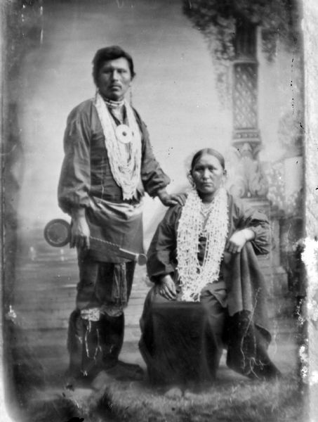 Copy photograph of a studio portrait of a Ho-Chunk man posing standing on the left in front of a painted backdrop. He is holding a war club, and is wearing regalia including several necklaces, a gorget, earrings, and garters. He has his left hand on the right shoulder of an older Ho-Chunk woman who is posing sitting on the right, and who is wearing several necklaces and earrings. Identified as Steve Waukon and his mother, the wife of Lou Waukon.