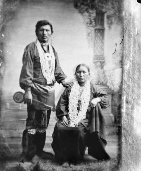 Copy photograph of a studio portrait of a Ho-Chunk man posing standing on the left in front of a painted backdrop. He is holding a war club, and is wearing regalia including several necklaces, a gorget, earrings, and garters. He has his left hand on the right shoulder of an older Ho-Chunk woman who is posing sitting on the right, and who is wearing several necklaces and earrings. Identified as Steve Waukon and his mother, the wife of Lou Waukon.