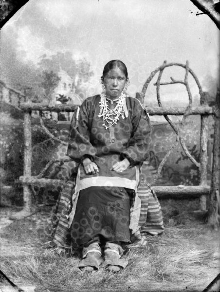 Studio portrait of a Ho-Chunk woman posing sitting in front of a prop wooden fence and a painted backdrop. She is wearing several necklaces, earrings, moccasins, and a ribbon work blouse, and is identified as Mabel White St. Cyr.