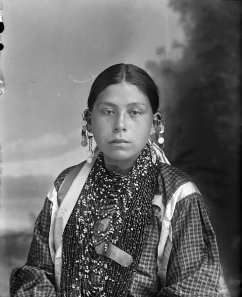 Waist-up studio portrait of a young Ho-Chunk woman posing sitting in front of a painted backdrop. She is wearing several necklaces, earrings, and streamers. She is identified as Joanna White Otter.