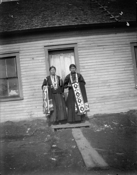 Two Ho-Chunk women posing standing outdoors in front of a doorway of a wooden building. They are wearing dark dresses and several necklaces, and holding ribbon work shawls. They are identified as Nellie and Sarah Windblowe, but not identified by position.