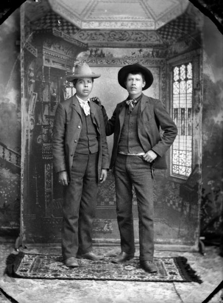 Studio full-length portrait of two Ho-Chunk men posing standing in front of a studio backdrop, and wearing suits and hats. The man on the right has his right hand on the left shoulder of the other man. They are identified as Dr. Bill Thunder standing on the left, and William Massey(Massie) standing on the right.
